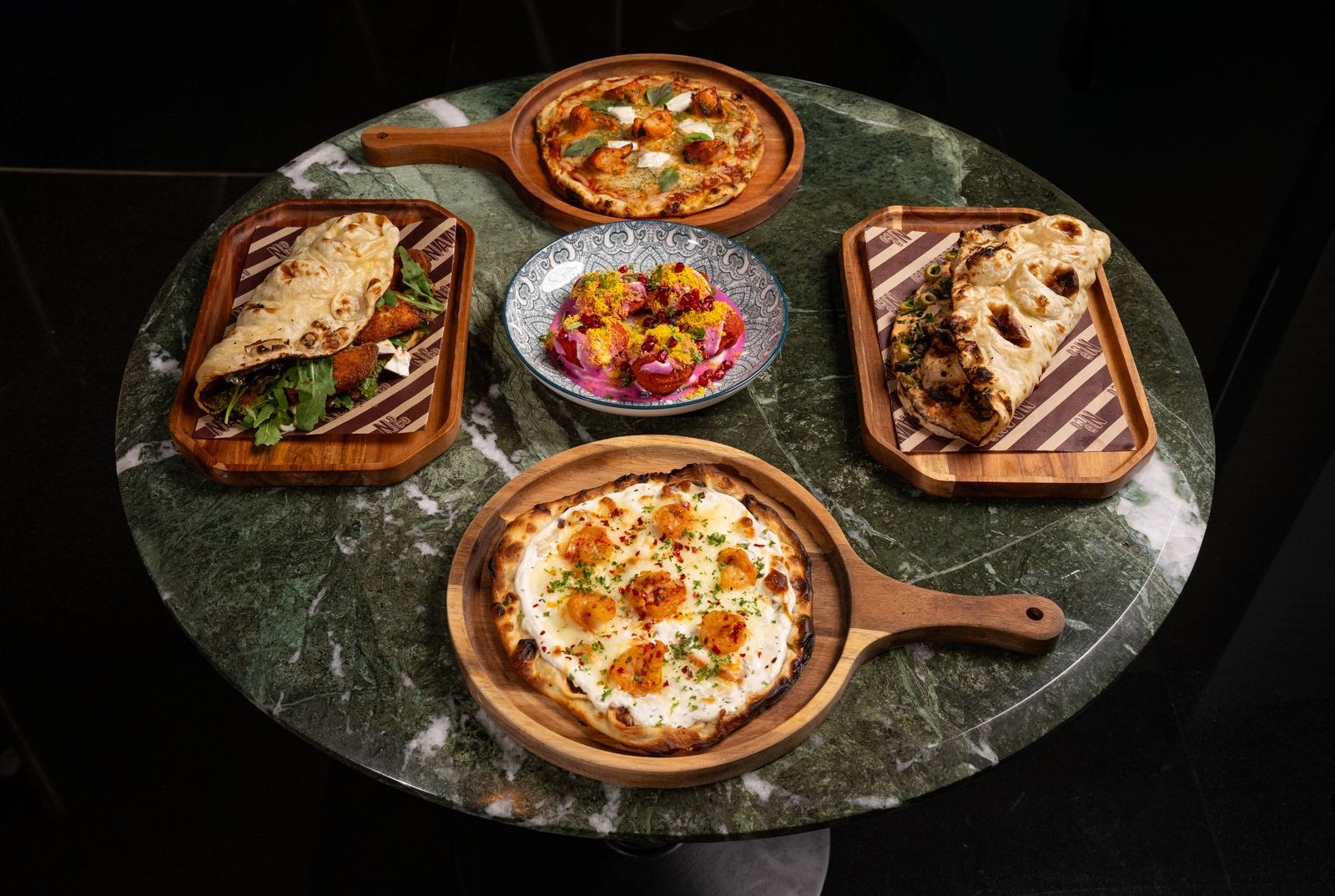 An overhead spread of Naan & Co dishes: naanwiches, pizzaans and a chaat, fired fresh and plated on wooden boards