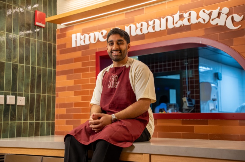 Ronit Bhatia in a Naan & Co apron, sitting on the counter beneath the Have a naantastic day sign